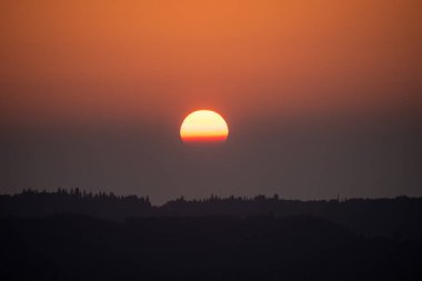 Güneş ufukta batıyor, turuncu ve kırmızı tonlarda sisli gökyüzünü boyuyor. Yuvarlanan tepelerin karanlık silueti ve Yunanistan 'da bir ormanın üzerinde güzel bir sonbahar günbatımı. Huzurlu doğa manzarası.
