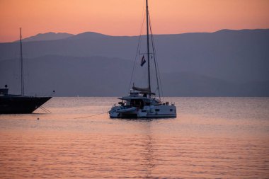 Alacakaranlıkta sakin, pembe renkli bir denize demirlemiş, ufukta puslu dağlar olan modern bir yelkenli katamaran. Yunan adalarında sakin bir tatil ve seyahat sahnesi