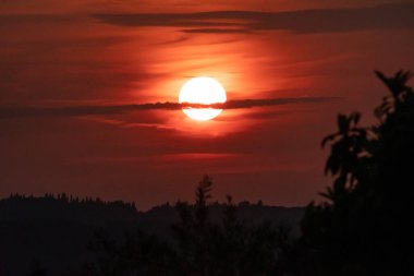 Güneş kırmızı ve turuncu bir alevle batar, ince ve karanlık bir bulut tarafından kısmen gizlenir. Kırsal Yunan manzarasının siluetinde dramatik ve güzel bir akşam sahnesi