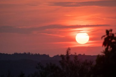 Parlak güneş diski yoğun bir şekilde parlayan dramatik, koyu kırmızı bir gün batımı. Alevli gökyüzü tepelerden ve ağaçlardan oluşan karanlık bir siluetin üzerinde asılı duruyor. Güçlü ve güzel bir gün sonu sahnesi yaratıyor.