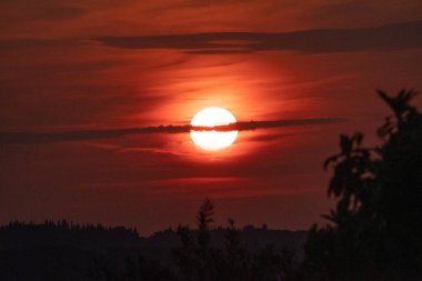 İnce bir bulutun ardında parlak güneşin battığı dramatik, koyu kırmızı bir gün batımı. Ateşli gökyüzü tepelerden ve ağaçlardan oluşan karanlık bir siluet üzerinde parlıyor, güçlü ve karamsar bir akşam manzarası yaratıyor.