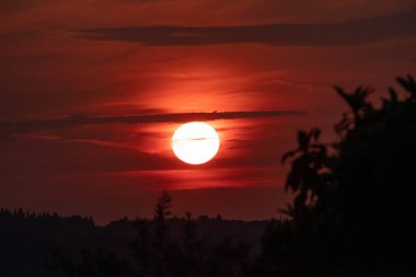 İnce bir bulutun ardında parlak güneşin battığı dramatik, koyu kırmızı bir gün batımı. Ateşli gökyüzü tepelerden ve ağaçlardan oluşan karanlık bir siluet üzerinde parlıyor, güçlü ve karamsar bir akşam manzarası yaratıyor.
