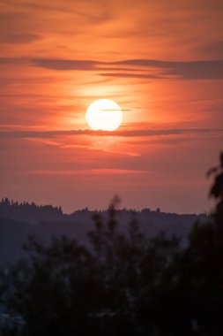 Parlak güneşin turuncu ve kırmızı gökyüzünde parlak bir parıltı oluşturduğu huzurlu ve güzel bir gün batımı. Ufukta tepenin karanlık silueti uzanıyor ve huzurlu bir akşam manzarası yaratıyor.