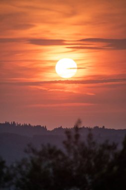Güzel bir günbatımının huzurlu, dikey manzarası. Güneş, uzak tepelerin karanlık siluetinin üzerinde, yumuşak turuncu bir gökyüzünde ışıl ışıl parlıyor. Huzurlu ve huzurlu bir hava yaratıyor.