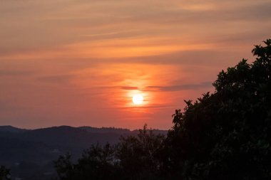 Yunan kırsalının yuvarlanan tepelerinin üzerinde güzel, sakin bir günbatımı. Sıcak turuncu ve kırmızı gökyüzü, karanlık bir ağaç silueti tarafından çerçevelenmiş huzurlu ve sakin bir akşam manzarası yaratır.