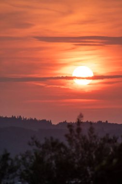 Huzurlu bir günbatımının güzel dikey kompozisyonu. Güneş, uzak tepelerin karanlık siluetinin ve Yunan kırsalındaki selvi ağaçlarının üzerinde yumuşak turuncu ve pembe bir gökyüzünde sıcacık bir şekilde parlıyor.