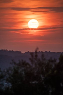 Dikey kompozisyonda güzel bir günbatımı. Parlak güneş, Yunan kırsalındaki tepelerin ve selvi ağaçlarının üzerinde sıcak turuncu ve pembe bir gökyüzünde asılı duruyor.
