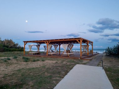 Alacakaranlıkta dolunay altında sallanan lüks gündüz yatakları olan romantik ahşap plaj çardağı. Deniz kıyısındaki bir otelde sakin ve büyülü bir gece sahnesi. Balayındakiler için mükemmel ya da okyanus kıyısından huzurlu bir kaçış.