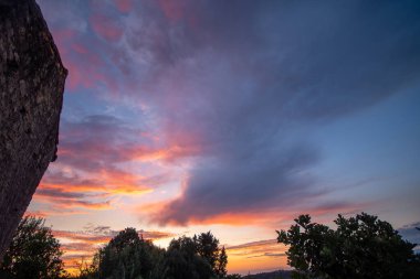 An awe-inspiring sunset view over rural Greece, with a vast, dramatic sky filled with textured clouds painted in brilliant shades of orange, pink, and deep blue