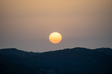 A soft, hazy sunset with a perfect orange sun orb suspended over dark blue hills. A serene and minimalist landscape capturing the quiet beauty of twilight