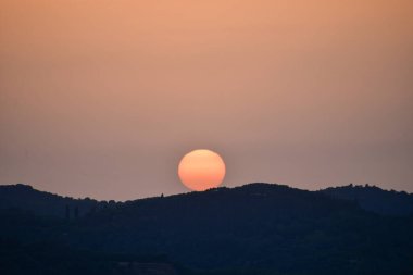 A picturesque sunset with a soft, pinkish-orange sun setting perfectly behind a silhouetted hill in a hazy sky, creating a peaceful and serene landscape
