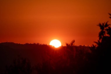 A brilliant sun sinks behind a silhouetted forest on the horizon, painting the sky in fiery shades of red and orange during a beautiful and dramatic sunset