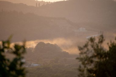 Ethereal morning mist and fog settle in a lush green valley during a golden hour sunrise, with a modern white villa visible on a distant hill. A hazy, atmospheric landscape in rural Greece