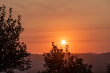 A breathtaking panoramic view of a golden sunset over rolling hills and mountains in the Greek countryside. The sky is painted with warm orange hues, framed by tree silhouettes for a scenic landscape