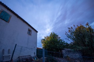A moody, wide-angle view of a rustic house at dusk, with a long-exposure sky filled with dramatic, streaky blue clouds. Tall reeds in the foreground add to the natural, rural atmosphere