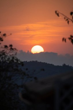 The sun sets in a soft orange sky over the rolling hills of Tuscany, Italy. A beautiful and serene evening landscape framed by blurred leaves, with a historic villa in the distance