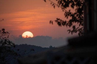 A tranquil sunset viewed from a balcony or window, framed by dark silhouettes of leaves. The golden sun sinks behind clouds and distant hills with a castle in a serene, picturesque landscape