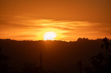The sun bursts with intense golden light as it sets behind silhouetted hills in Greece. A powerful and vibrant sunset scene, painting the entire sky a brilliant shade of orange