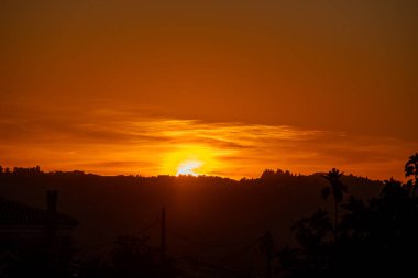 A mesmerizing sunset over the Greek hills, with the fiery orange sun sinking below the horizon. The warm, glowing sky creates a beautiful silhouette of the tranquil landscape