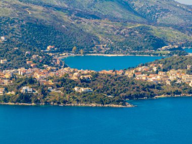 Yunan adası Corfu 'daki Kassiopi' nin sakin körfezinin havadan görünüşü, kıyıya kurulmuş büyüleyici köy ve canlı mavi suyu çevreleyen yemyeşil tepeler.
