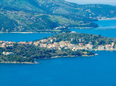 Yunan adası Corfu 'daki Kassiopi' nin sakin körfezinin havadan görünüşü, kıyıya kurulmuş büyüleyici köy ve canlı mavi suyu çevreleyen yemyeşil tepeler.