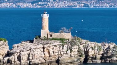 Hava aracı görüntüleri engebeli, kayalık bir adadaki tarihi deniz fenerine yaklaşıyor. Uzak bir deniz fenerinin sinematik görüntüsü derin mavi denize ve uzak Saranda şehrine karşı