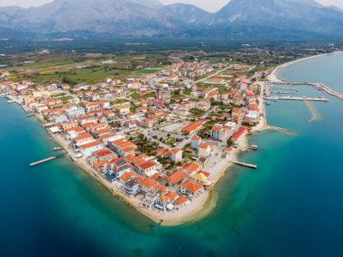 Yunanistan 'ın Mytikas kıyı köyünün üzerinde uçan yüksek hava aracı görüntüsü. Deniz kenarı bir yarımadada, marinası, kırmızı çatılı evleri ve turkuaz İyon denizi var..