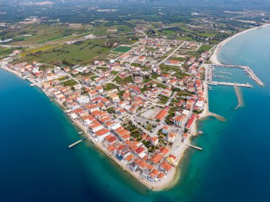 Başlık: Kırmızı çatıları, tekne marinası ve turkuaz Akdeniz 'de uzun kumlu bir plajı olan kıyı Yunan köyünün geniş hava manzarası.