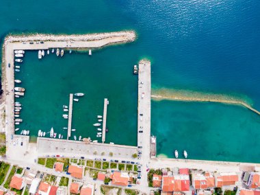 Rıhtımlı bir sürü teknesi, uzun bir iskelesi ve turkuaz deniz kıyısında kırmızı çatılı evleri olan bir sahil köyünün havadan görünüşü..