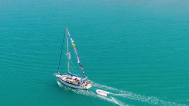 Yüksek açılı insansız hava aracı videosu bayraklı bir yatı takip ediyor, canlı bir turkuaz okyanusu geçen bir botu çekiyor.