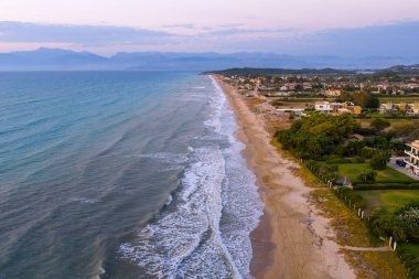 Korfu, Yunanistan 'da uzun kumlu bir plajın eril insansız hava aracı manzarası. Sahil tatil beldeleri ve evler kıyı boyunca uzanırken, İyon Denizi Yunanistan 'ın uzak dağlarıyla buluşuyor