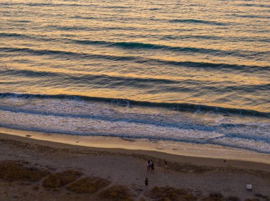 Gün batımında kumlu bir plajda yürüyen insanların yukarıdan aşağı görüntüsü. Altın ışık, denizin yumuşak dalgalarını yansıtarak huzurlu bir akşam atmosferi yaratır.