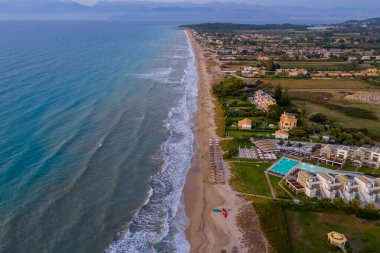 Yunanistan 'ın Korfu kentindeki lüks bir sahil beldesinin çarpıcı hava manzarası. Büyük bir yüzme havuzu, modern binalar ve güneşlikler turkuaz İyon Denizi kıyısında uzun ve kumlu bir sahilde bulunur.