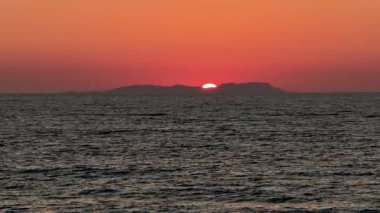 Uzak bir adanın siluetinin arkasında, muhtemelen Diapontia Adaları 'nın ardında, karanlık ve dalgalı İyon Denizi' nin üzerinde canlı kırmızı bir güneşin batışının yakın çekim görüntüsü. Büyüleyici Corfu günbatımı