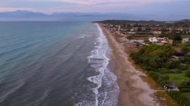 Yunanistan 'ın Corfu kentindeki manzaralı kumlu bir sahilde hava aracı görüntüleri. Turkuaz dalgalar, sahil evleri ve tatil beldeleri ve güzel bir alacakaranlık gökyüzünün altındaki uzak dağları gösteriyor.