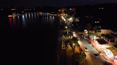 Ipsos 'un gece drone görüntüsü, Korfu kıyı şeridi. Aydınlatılmış ana yol boyunca uçmak, sokak lambalarının, araba farlarının ve deniz kıyısındaki işletmelerin parlaklığını yakalamak.