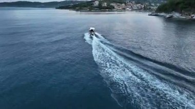 Karanlık çöktüğünde kıyı şeridi boyunca fermuar çeken sürat teknesi Derin Mavi Deniz 'de beyaz bir uyanış, Uzakta Sahil Köyü' nün havadan görünüşü