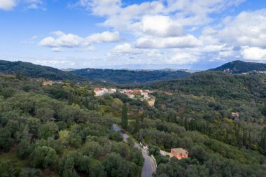Korfu adasındaki yeşil tepelere ve zeytinliklere kurulmuş geleneksel bir Yunan köyünün panoramik hava manzarası mavi bulutlu bir gökyüzünün altında