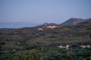 Kuzey Korfu 'nun geniş panoramik manzarası, Yunanistan' da yumuşak mavi ve mor bir akşam gökyüzünün altında uzanan geniş bir zeytin ağaçları ormanıyla çevrili tepedeki Nimfes köyünü kapsıyor..