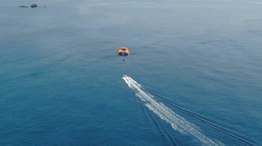 Hız teknesinin havadan görüntüsü renkli paraşütle çekiliyor ve güneşli bir yaz gününde derin mavi deniz suyunda paraşütle atlayan biri var.