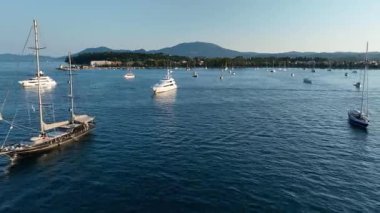 Güneşli bir yaz günü kıyı kenti yakınlarındaki sakin bir mavi deniz koyuna demirlemiş lüks yatların ve yelkenlilerin insansız hava aracı görüntüsü.