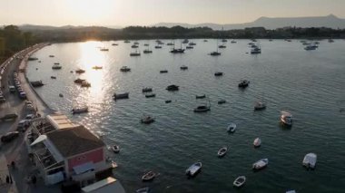 Gün batımının güzel görüntüsü Corfu 'nun marinasının sakin sularını yansıtıyor. Sahile yakın kıyıda demirlemiş sayısız küçük tekne ve kasabanın silueti yer alıyor.