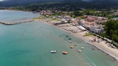 Yunanistan 'ın Korfu kentindeki hareketli bir sahil ve kıyı kasabasının canlı hava manzarası, renkli tekneler, sayısız güneşli yataklar ve parlak bir yaz gününde açık Akdeniz sularının keyfini çıkaran insanlar.