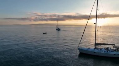 Günbatımında demir atmış lüks bir yelkenlinin yakın çekim görüntüsü. Mürettebat güvertede dinleniyor, deniz sakin ve gökyüzünde yumuşak bulutlar var. İyon Denizi yelkenli tatili