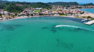 Yunanistan 'ın Corfu kentindeki bir sahil kenti yakınlarındaki kristal berrak turkuaz sularda daire çizen bir sürat teknesinin insansız hava aracı görüntüsü. Canlı yaz tatili sahnesi