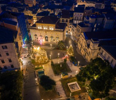 Yunanistan 'ın Corfu Town kentindeki San Giacomo Meydanı' nın panoramik gece manzarası, belediye binasını, Katolik Katedrali 'ni ve yukarıdan neşeli Noel piyasa süslemelerini ele geçiriyor.