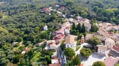 Yunanistan 'ın Korfu kentinde, çan kulesi, kırmızı çatılı evler ve yemyeşil ormanlarla çevrili dar sokakları olan geleneksel bir kilisenin hava aracı görüntüleri..
