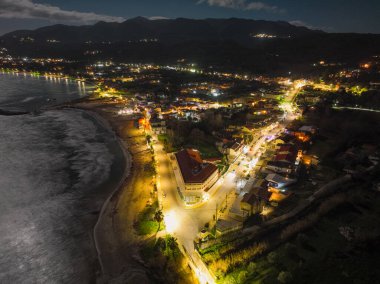 Korfu Yunanistan 'da Roda plajı ve kıyı şeridi gece görüşü, aydınlatılmış sahil köy tatil beldesi, kıyıda dalgalar kırılıyor ve karanlık bir gökyüzü altında uzak dağlar