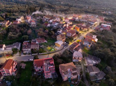 Korfu Yunanistan 'ın Xanthates köyünün hava manzarasında alacakaranlıkta duman tüten bacaları ve aydınlatılmış caddeleri olan geleneksel evler görülüyor
