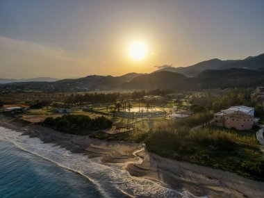 Yunanistan 'ın Kuzey Korfu kentindeki dalgalı dalgalar, altın ışık ve uzak dağlarla birlikte Acharavi sahilinin uzun kumlu kıyı şeridi üzerinde gün batımı manzarası